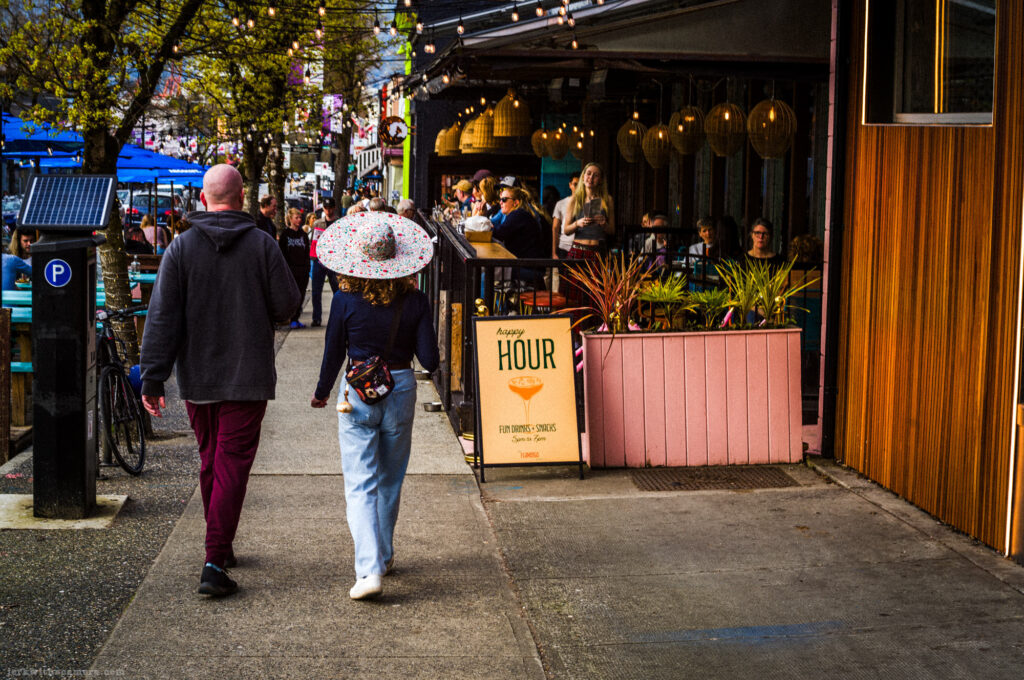 A couple walks past a restaurant patio with a happy hour sign on the sidewalk.