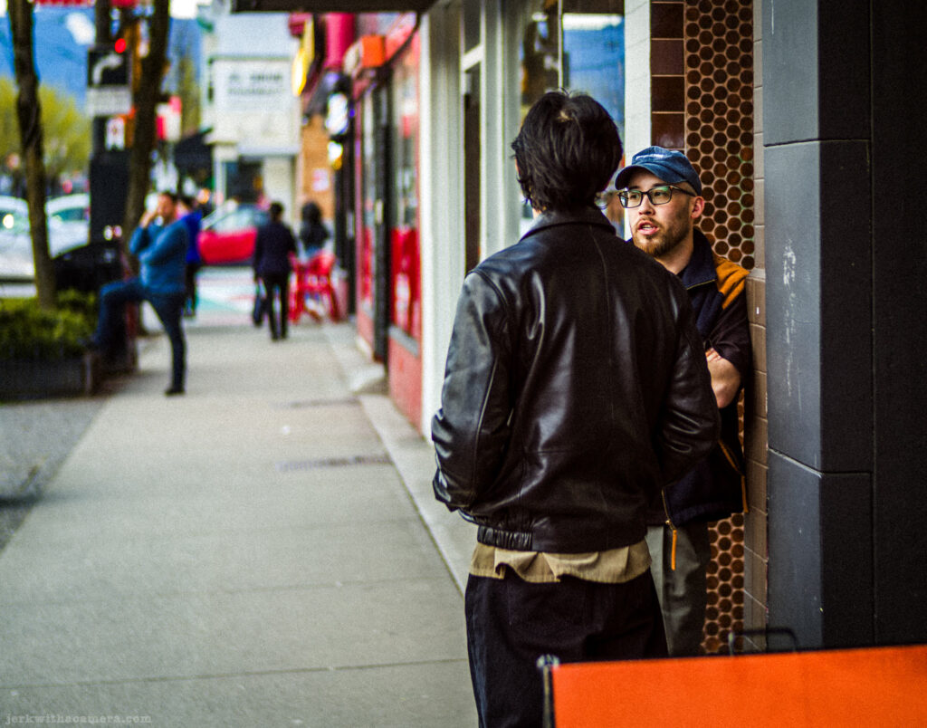 Two men stand talking outside a storefront on a busy sidewalk.