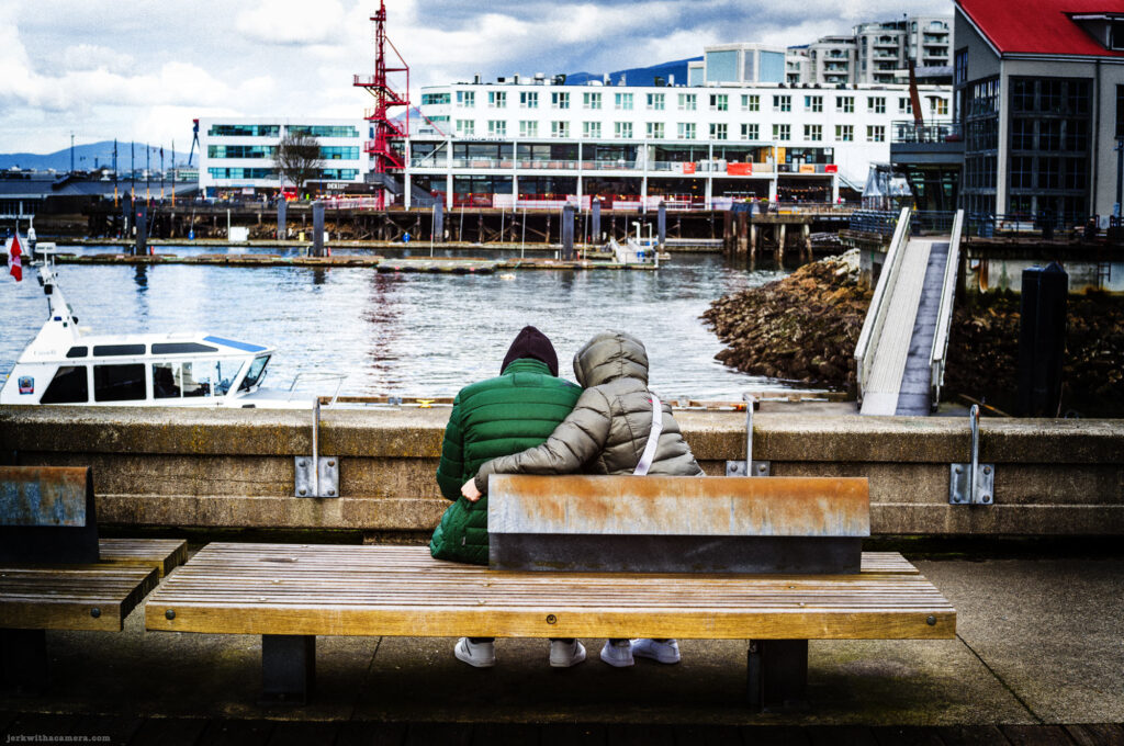 Two people in winter jackets sit on a waterfront bench with their backs to the camera.