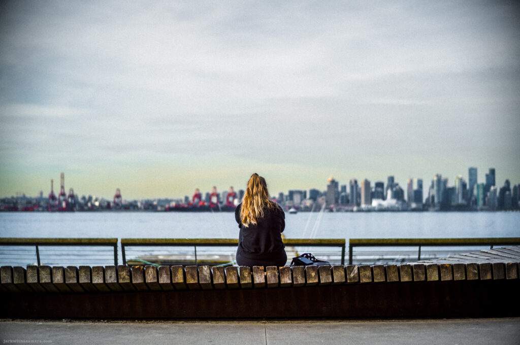 Rear view of a woman with long blonde hair sitting on a curved bench by the water, facing the Vancouver skyline across Burrard Inlet
