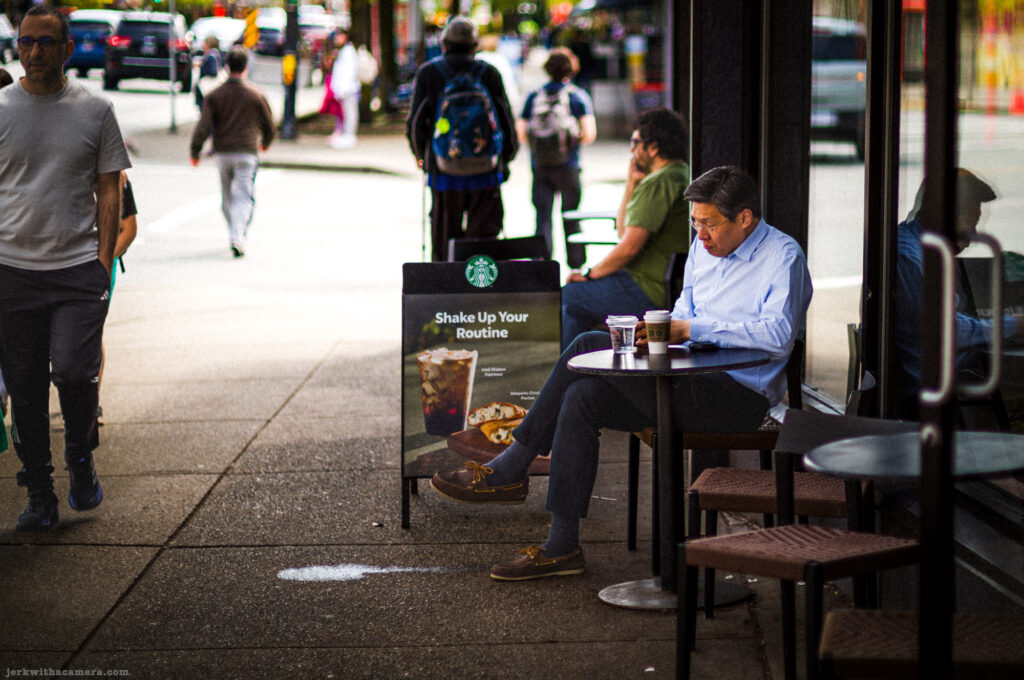 Man sitting outside a Starbucks on a city sidewalk reading with a drink beside him
