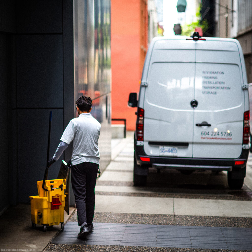 A man with cleaning equipment walks past a van in an alley