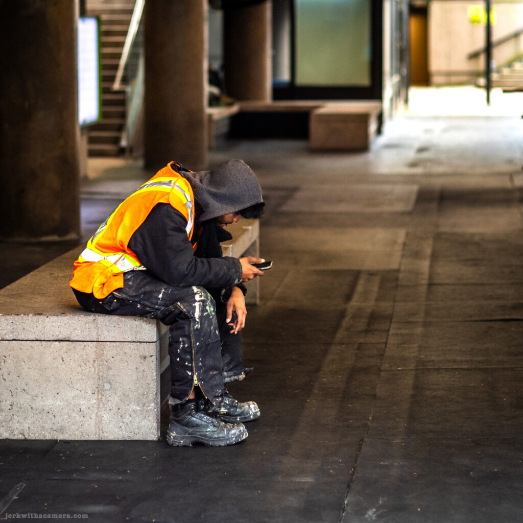 Worker in a reflective vest sits on a concrete bench looking at phone