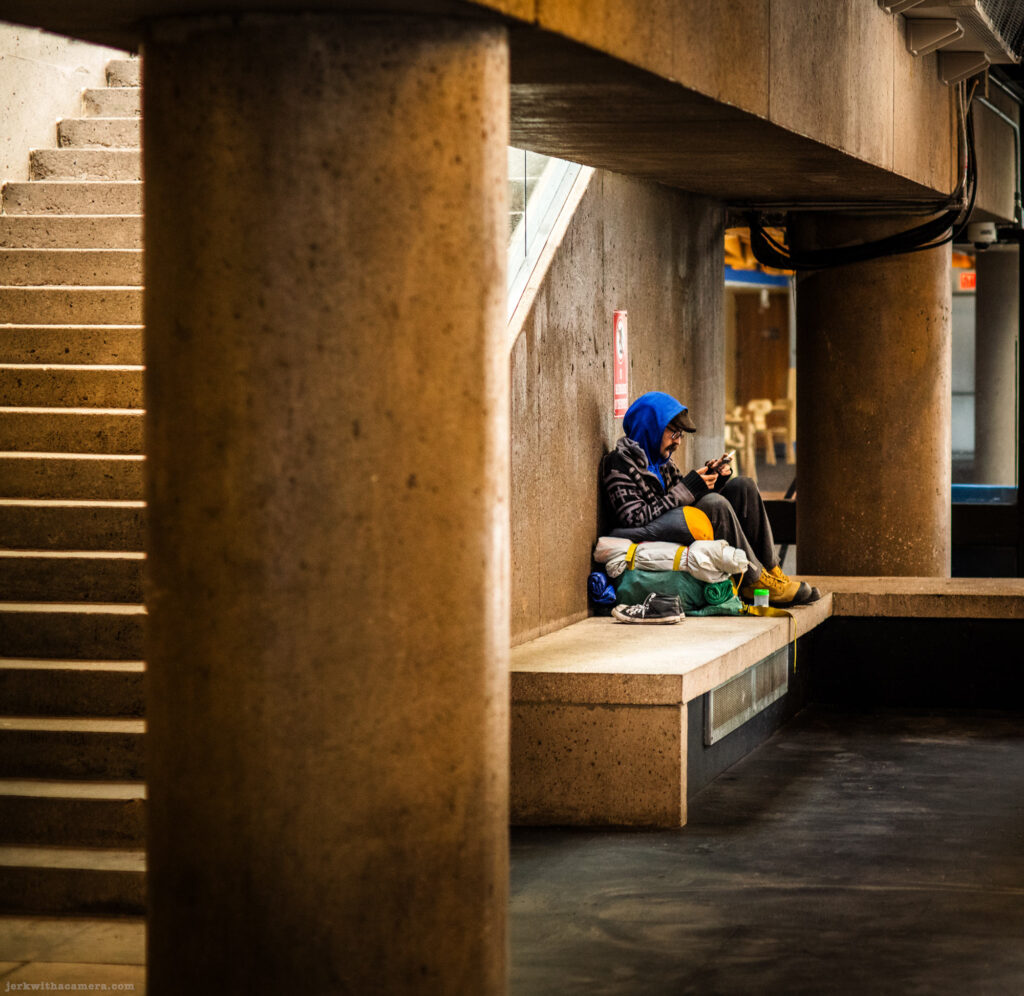 Person in blue hoodie seated on sleeping gear under concrete structure