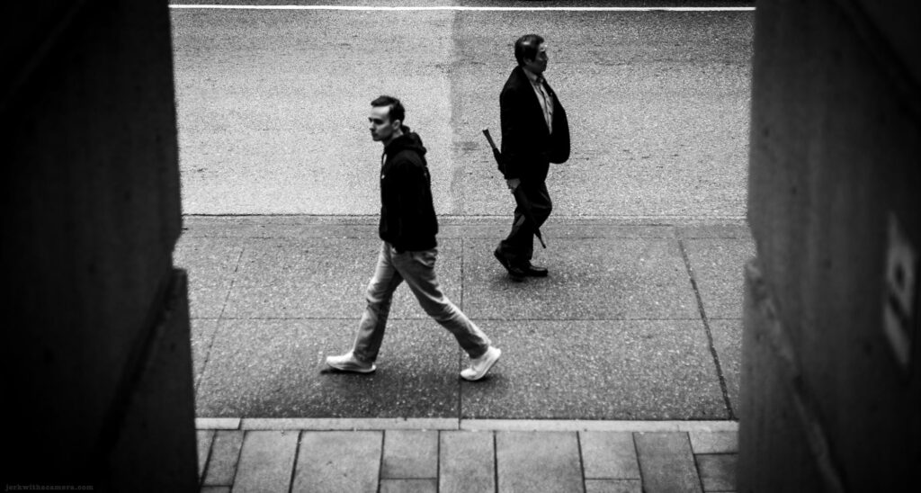 Two men walking in opposite directions on a sidewalk, black and white