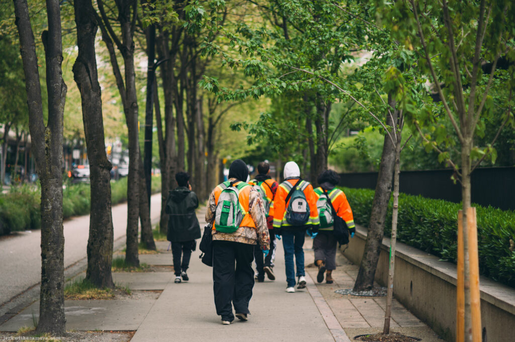 Group of workers in orange safety vests walking down a tree-lined sidewalk