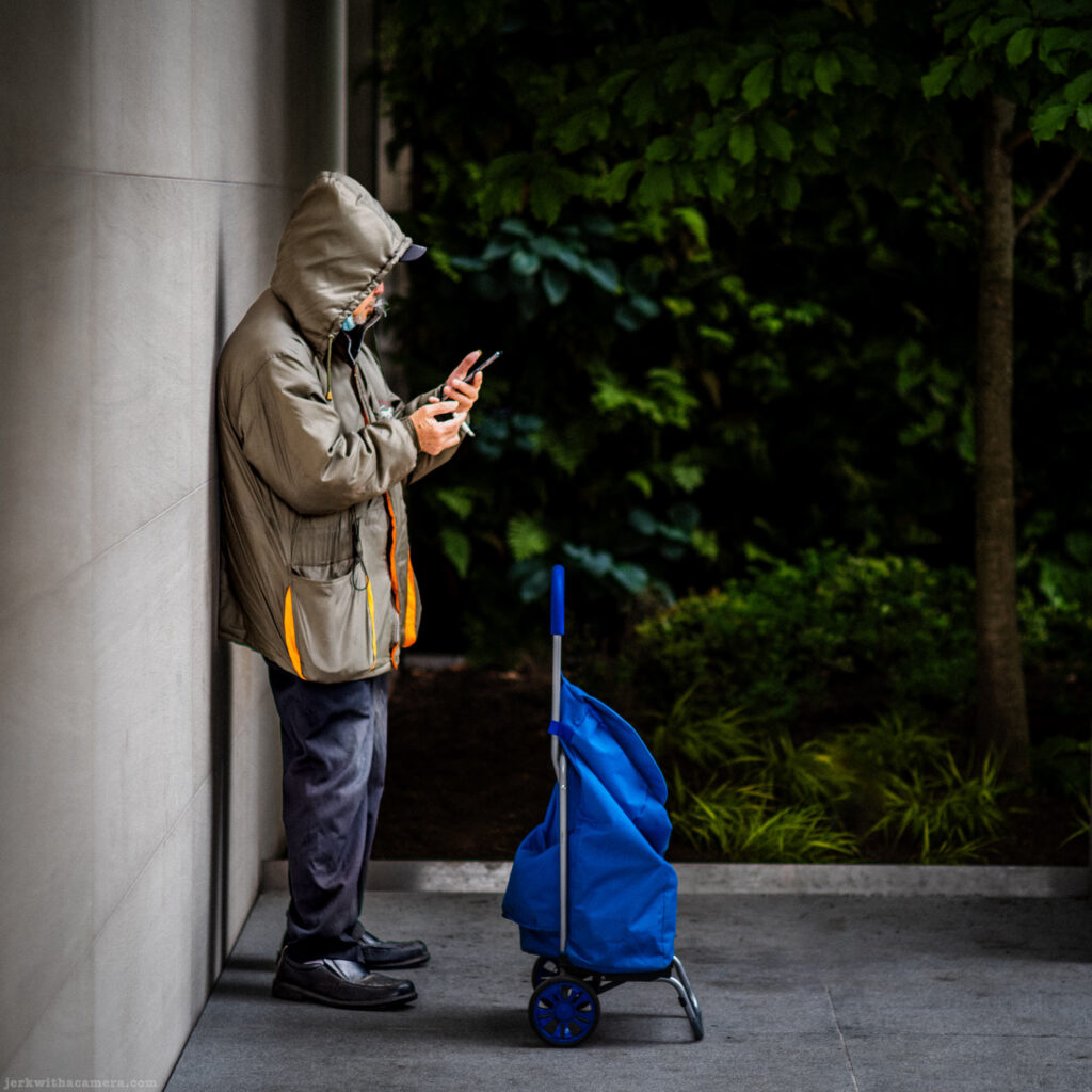 Man in hooded coat looking at phone beside a blue wheeled cart