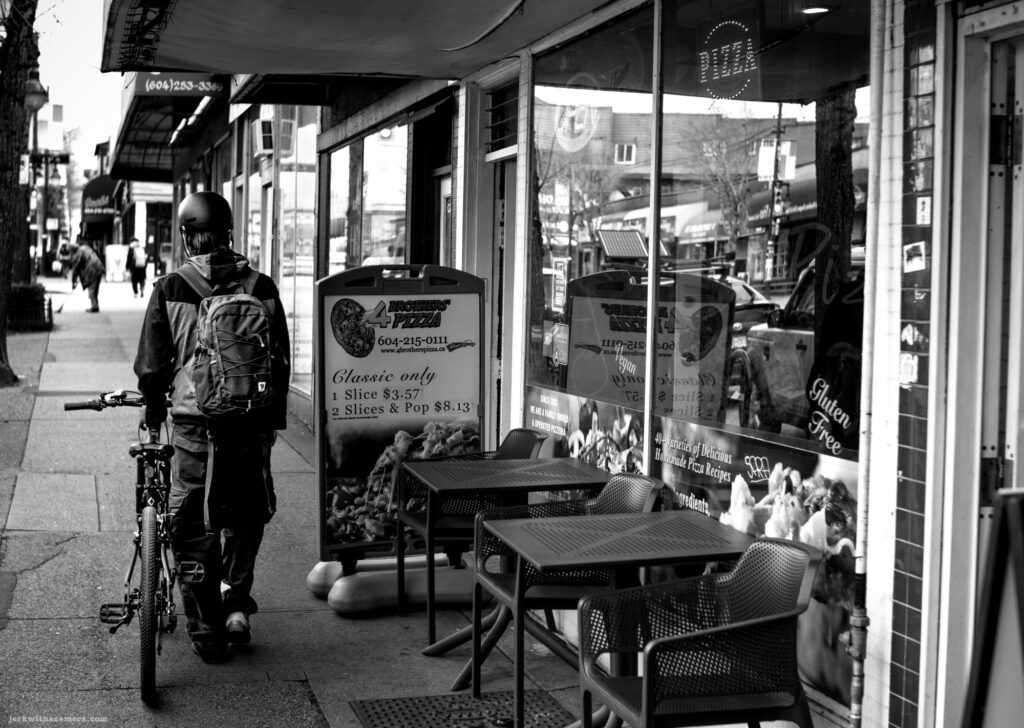 A person with a bicycle standing outside a pizza shop with a sidewalk sign