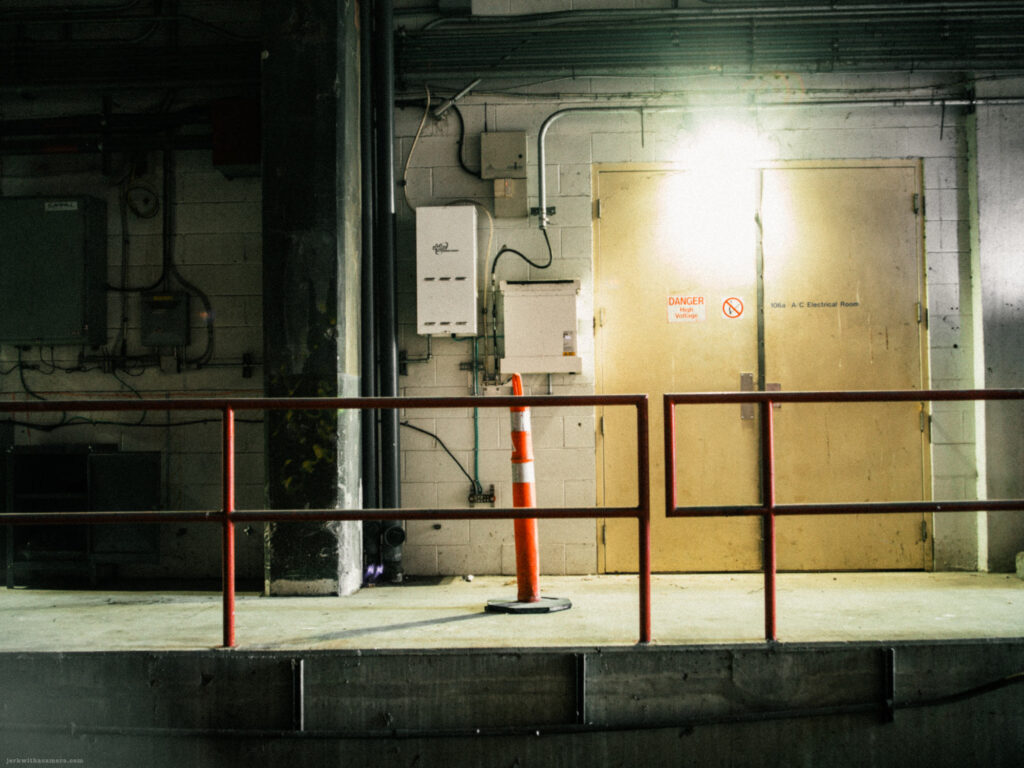 Industrial hallway with a closed metal door and overhead pipes lit by a single light