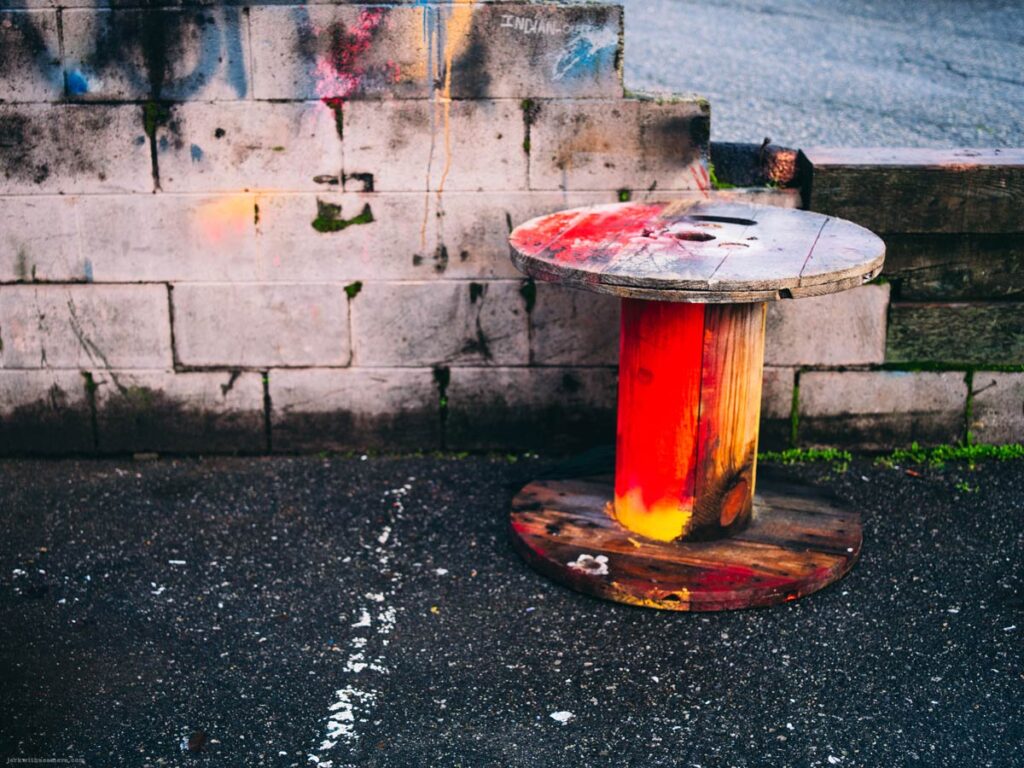 A wooden cable spool painted red and yellow placed on asphalt in front of a concrete block wall.