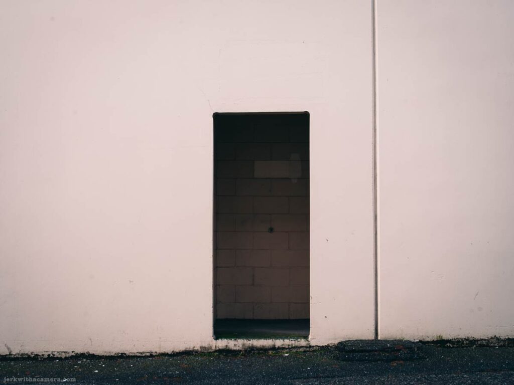 A narrow open doorway in a plain exterior wall leading into a dark interior space.