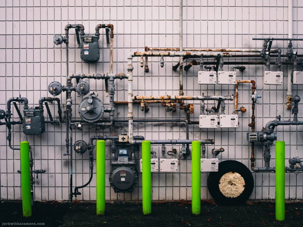 Gas meters and exposed piping mounted on a tiled exterior wall with green bollards in front at Lonsdale Quay in North Vancouver
