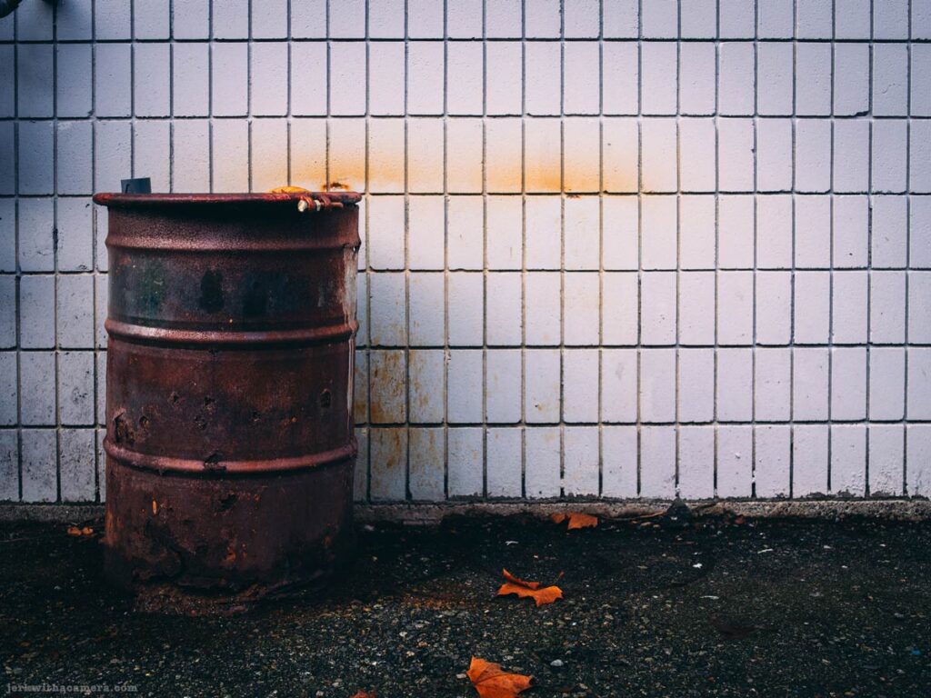 Rusty metal barrel against a white tiled wall with fallen leaves on the ground in North Vancouver