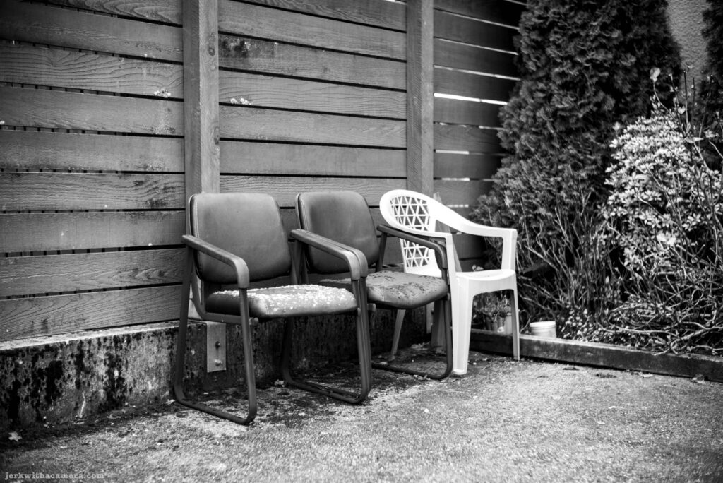 Two padded office chairs and one white plastic patio chair placed against a wooden fence beside shrubs