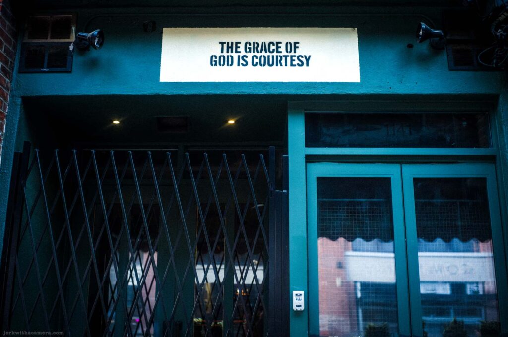 Illuminated sign reading The Grace of God is Courtesy above gated entrance