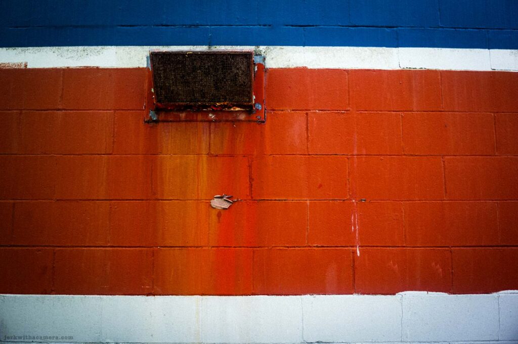 Red, white, and blue painted brick wall with rusted vent