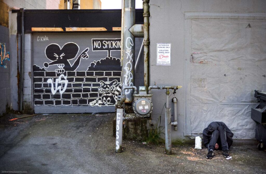 Graffiti on garage door beside utility pipe and person sitting against wall in alley