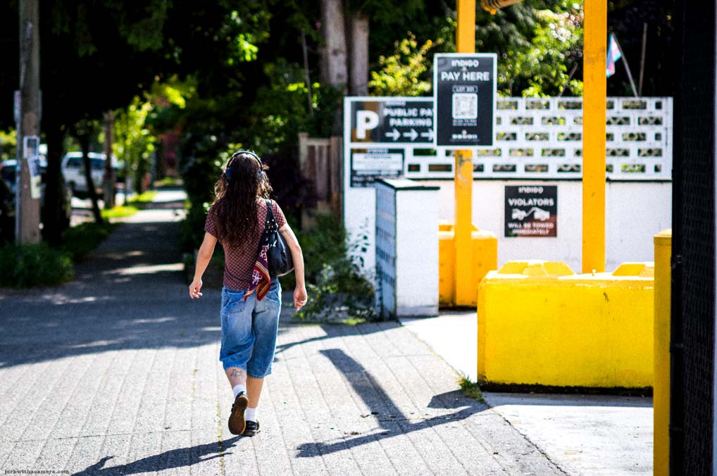 Person walking down a sidewalk past yellow parking barriers and signs