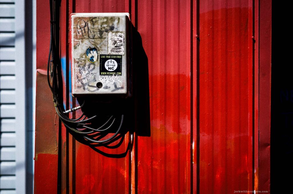 Sticker-covered electrical box mounted on a red corrugated wall