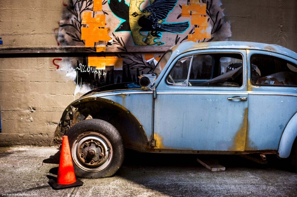 Old blue car parked beside a wall with graffiti and mural art