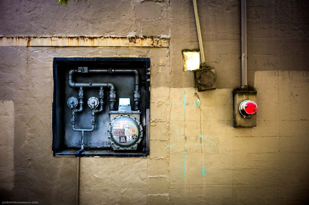 Gas meter, pipe, speaker, and red button mounted on concrete wall on Commercial Drive in Vancouver