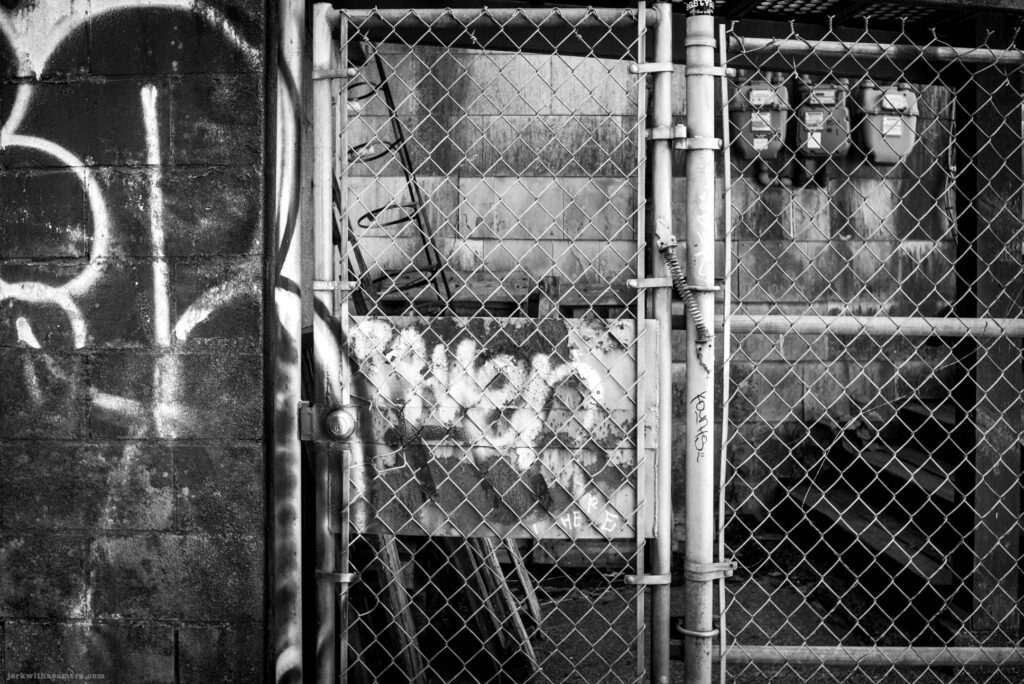 Chain-link fence with graffiti, plywood, and utility meter on Commercial Drive in Vancouver