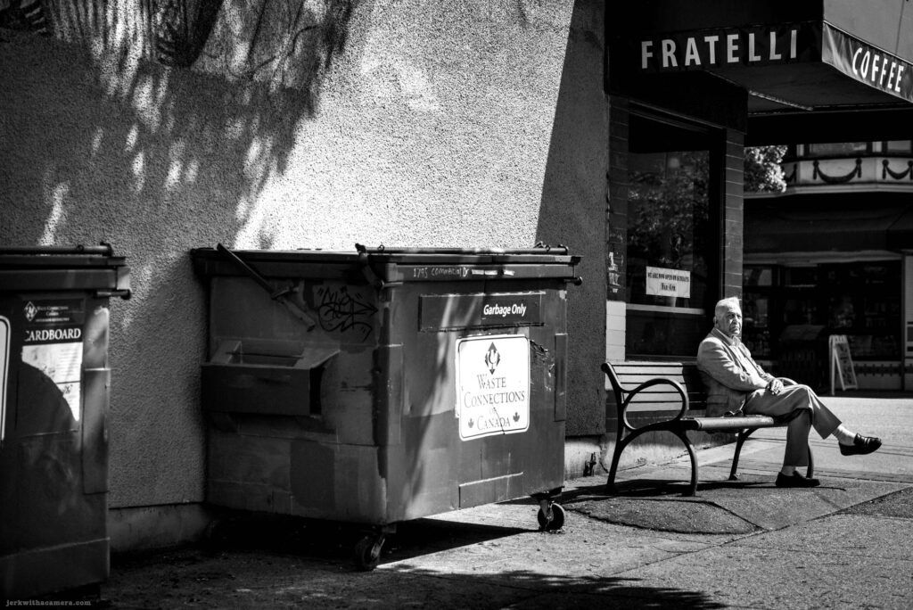 Man sitting on bench beside dumpster outside coffee shop