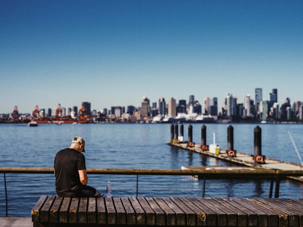 Person seated on a wooden platform beside the waterfront with a city skyline in the distance.