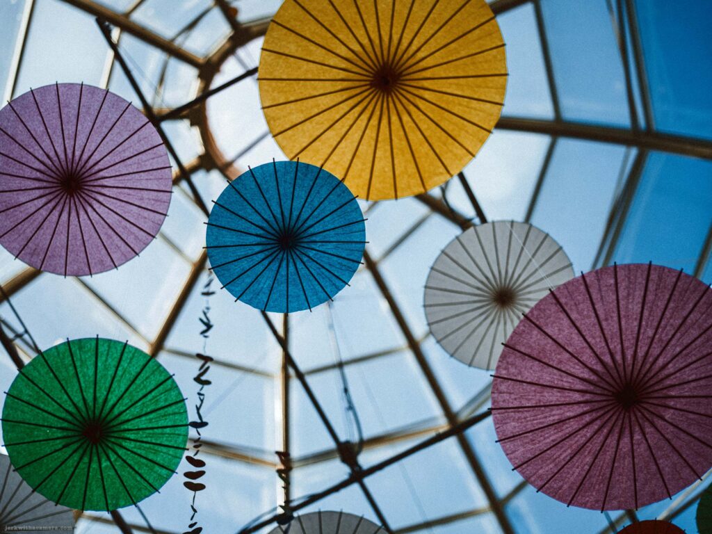 Coloured paper umbrellas suspended beneath a glass roof structure with sunlight shining through.