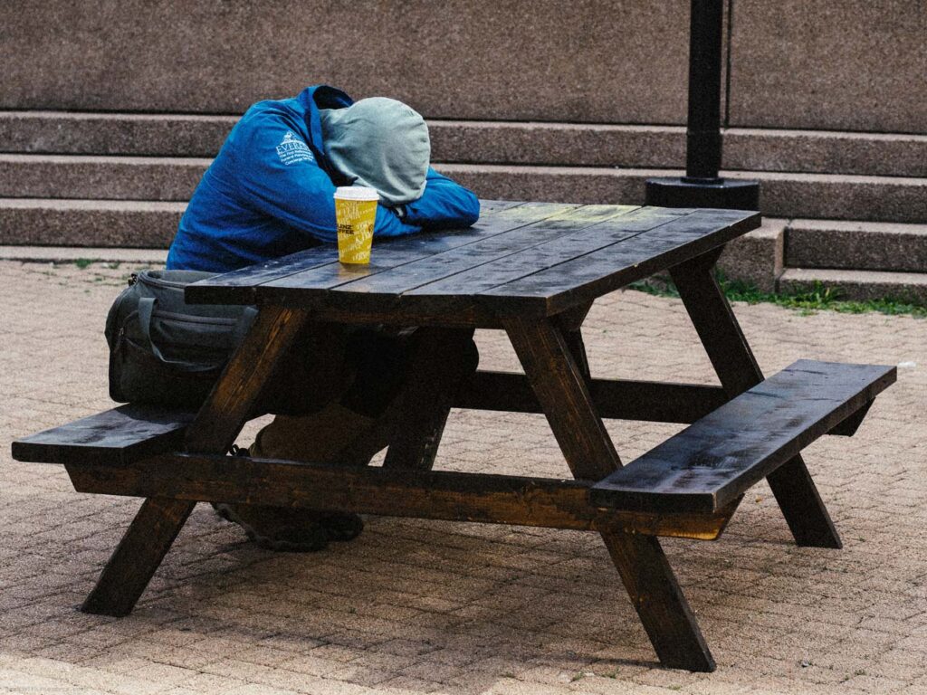 Person in blue jacket slumped over a dark wooden picnic table in a public plaza. Yellow cup on table. Pedestrians and cars in background.