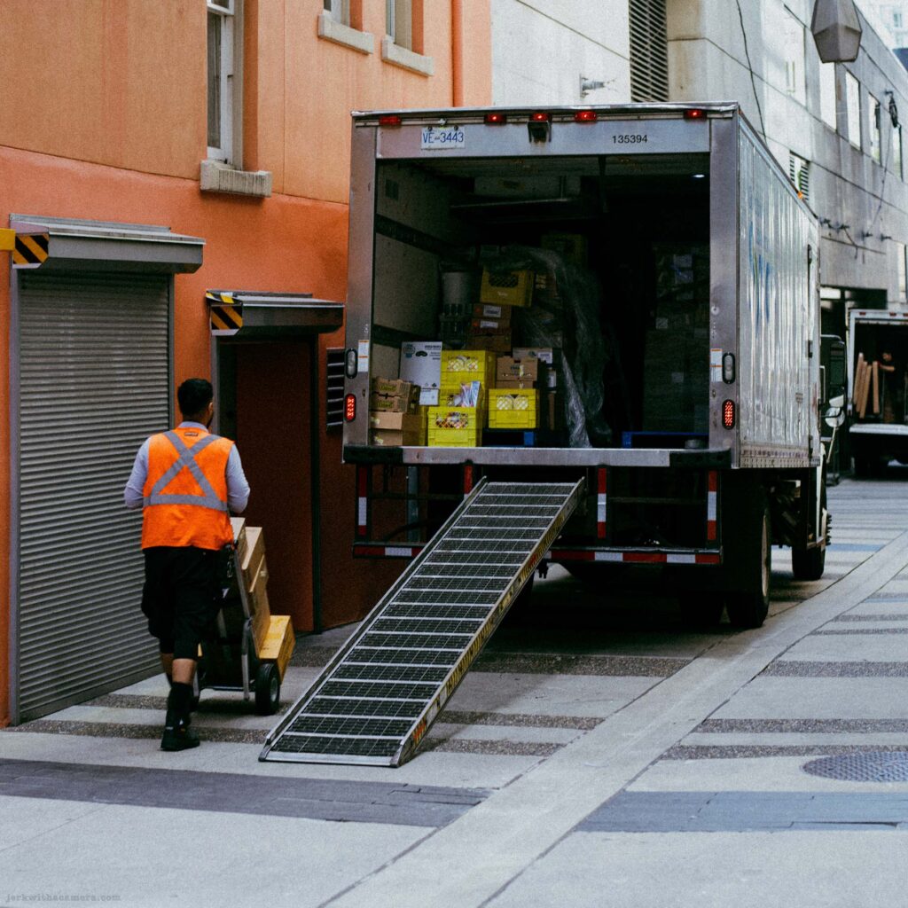 Delivery worker in orange vest unloading cardboard boxes from a grey truck via a loading ramp in a downtown alley. Second truck visible in background.