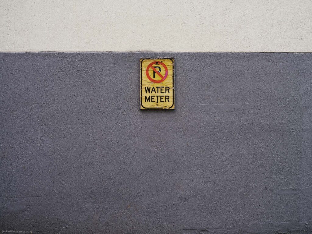 Weathered yellow no-parking sign reading Water Meter centred on a wall painted grey below and white above.