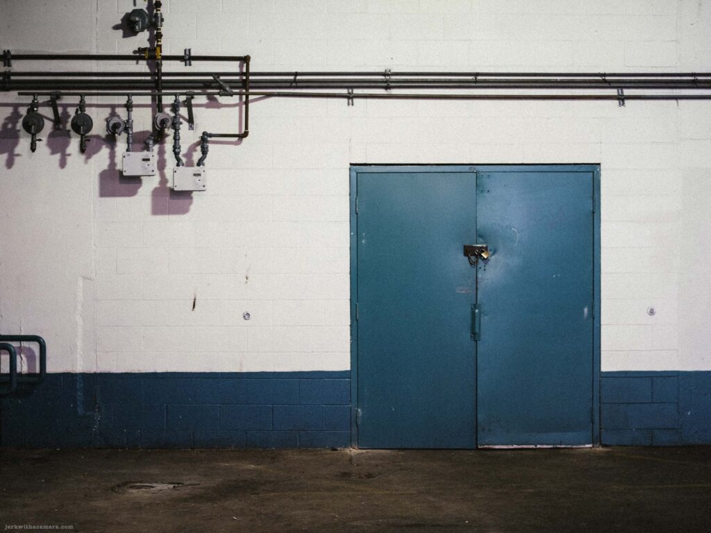 Green double door in a white concrete block wall with gas meters and piping mounted on the wall to the upper left.