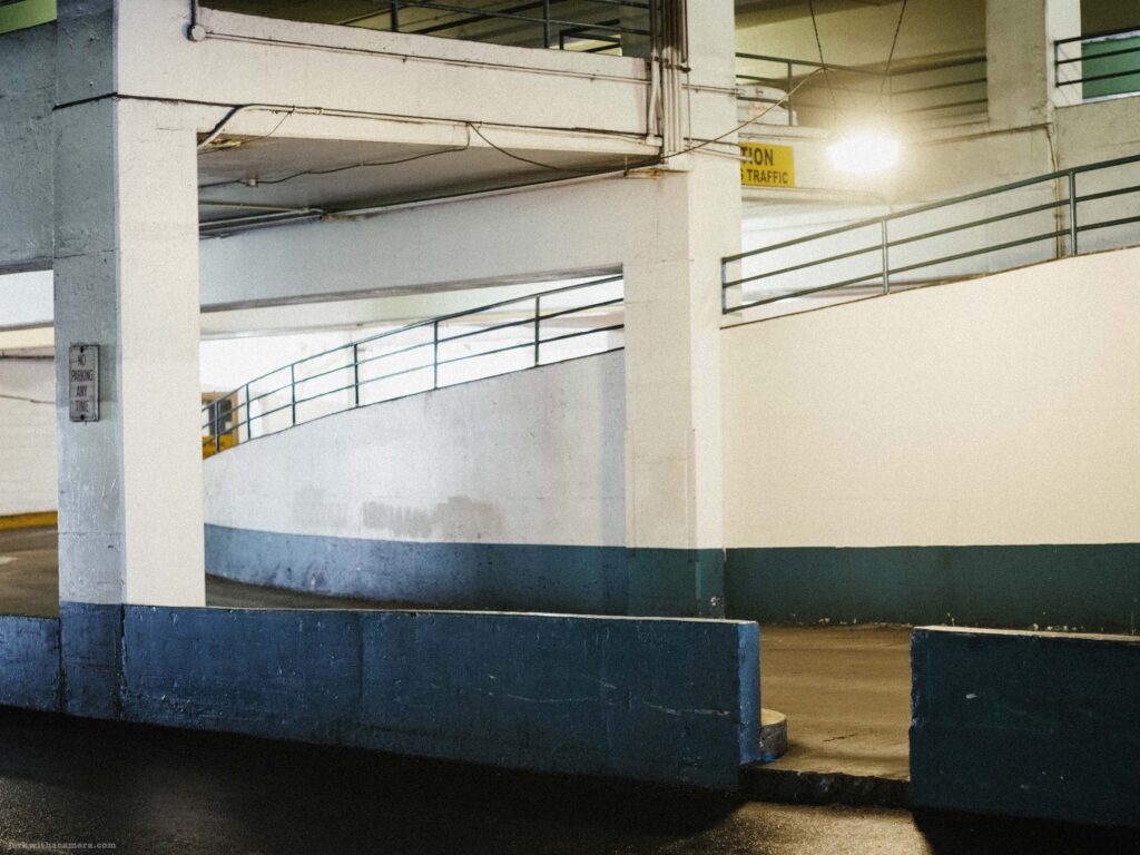 Parking garage interior with curved spiral ramp, concrete column, metal railings, and partial Caution Traffic sign at upper right.