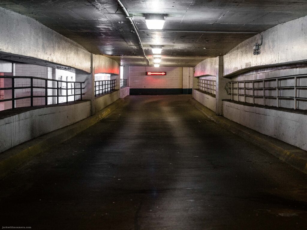 Empty parking garage drive lane at night. Fluorescent ceiling lights recede into the distance. Red neon directional arrow sign on the right wall.