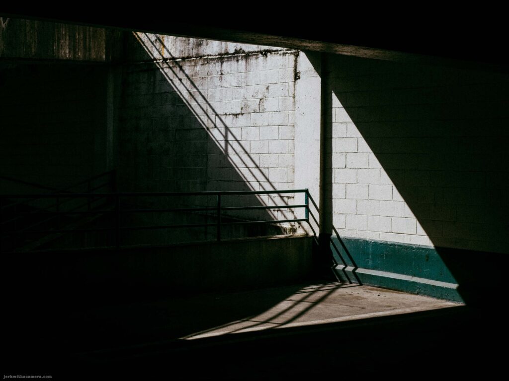 Wider view of parking garage stairwell corner with diagonal sunlight shaft, metal railings, concrete block wall, and teal lower band.