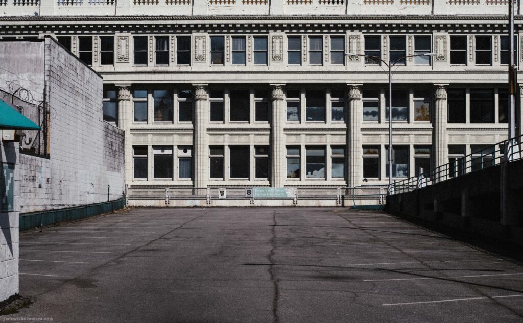 Wider view of neoclassical building facade with white brick wall at left and parking ramp structure at right. Empty paved lot in foreground.