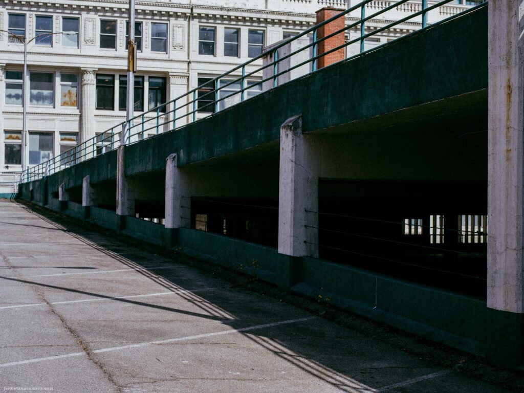 Multi-level parking ramp with teal painted edges and white columns beside it. Neoclassical building visible behind.