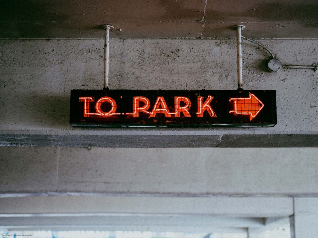 Red neon TO PARK sign at darkest exposure. Sign glow is most vivid. Concrete ceiling nearly black. Light from below visible at bottom.