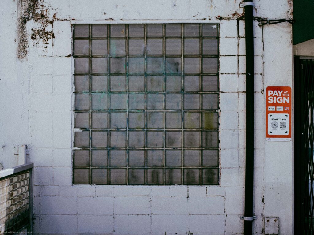 Wider view of glass block grid on white concrete block wall. Black drainpipe and Pay and Sign sign at right. More of the wall visible.