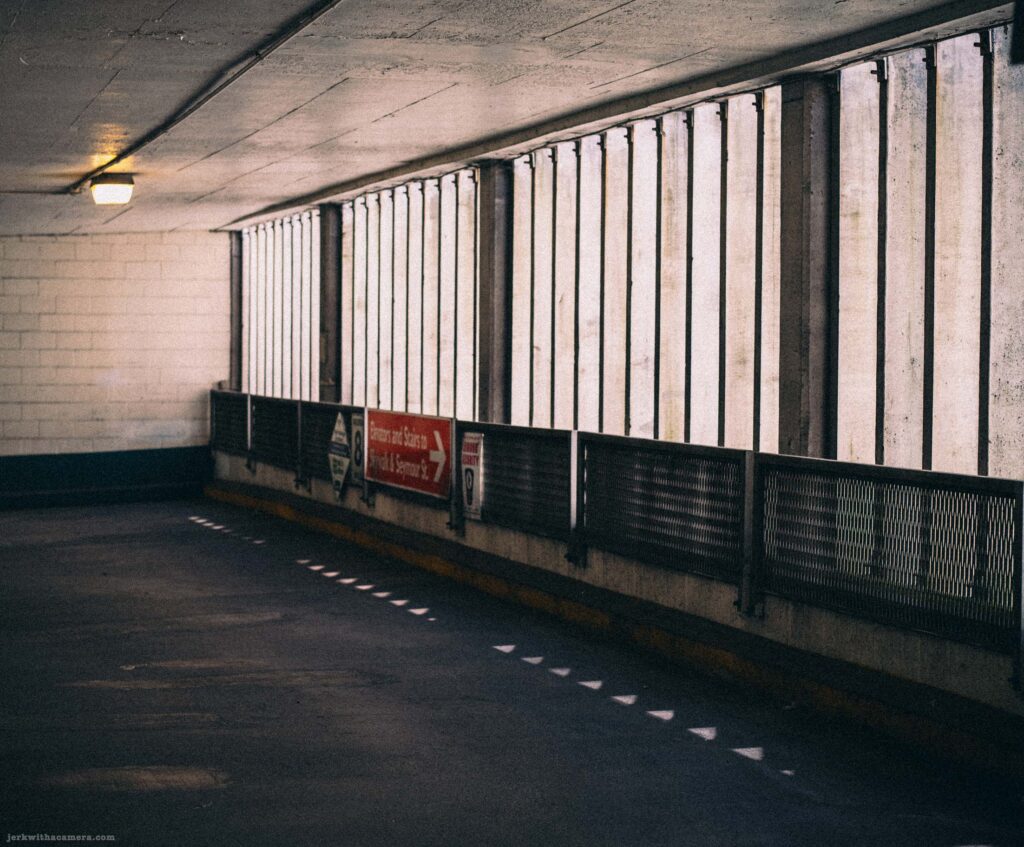 Parking garage interior lane with metal louvre panels above a railing, a pendant light, a teal lower wall band, and an orange directional sign.