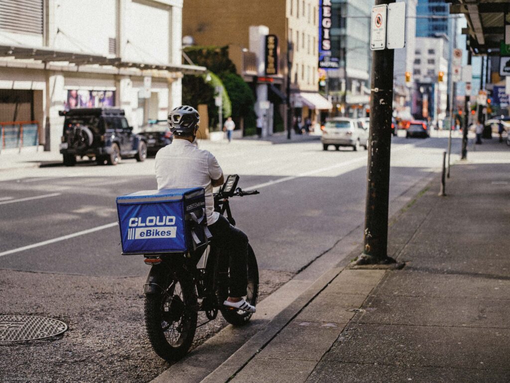 Delivery rider on Cloud eBikes cargo bike at curb, better exposed. Blue delivery box visible. Downtown street and Regis Hotel sign in background.