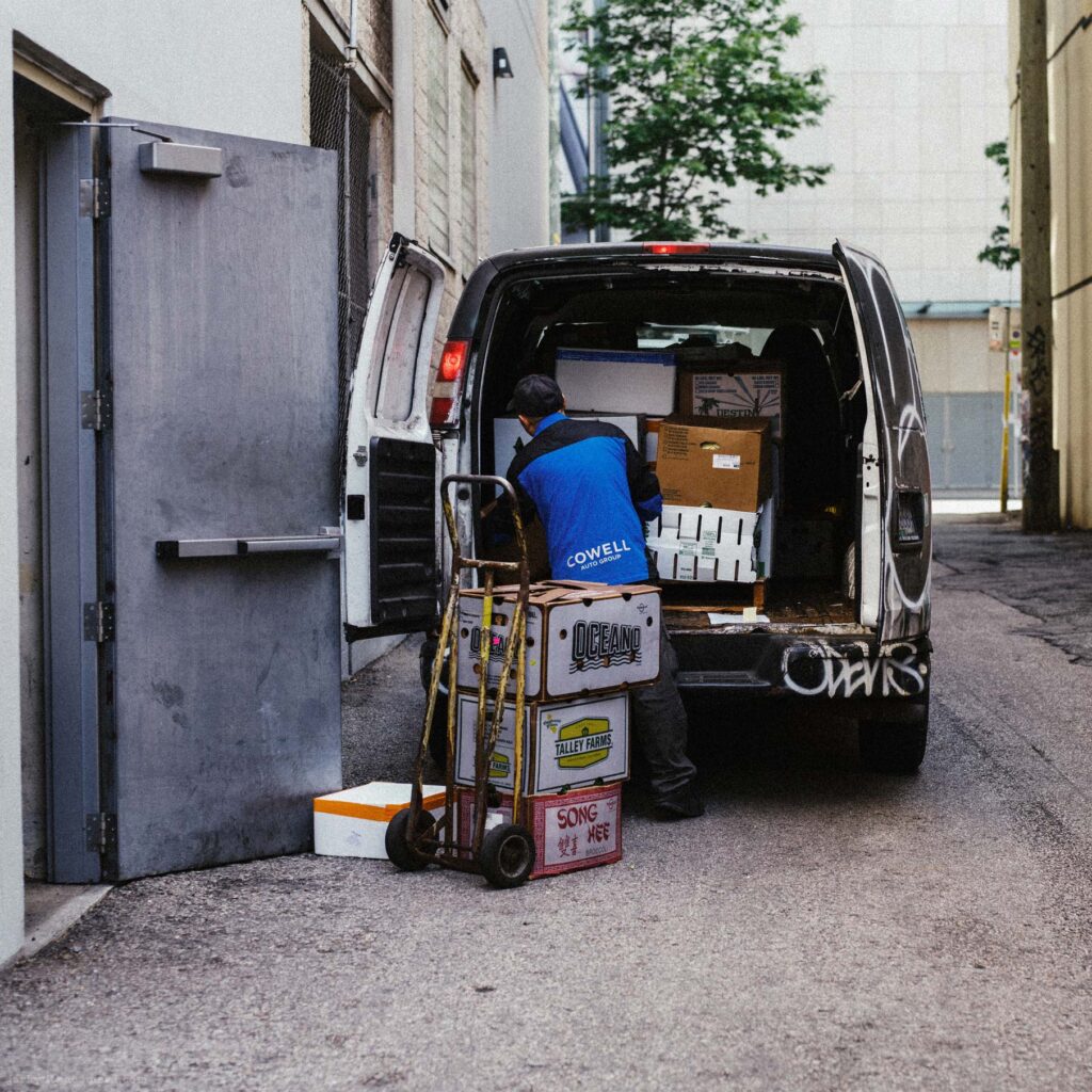 Delivery worker in blue uniform loading a white cargo van with cardboard boxes from a hand truck. Grey door and orange cone visible.