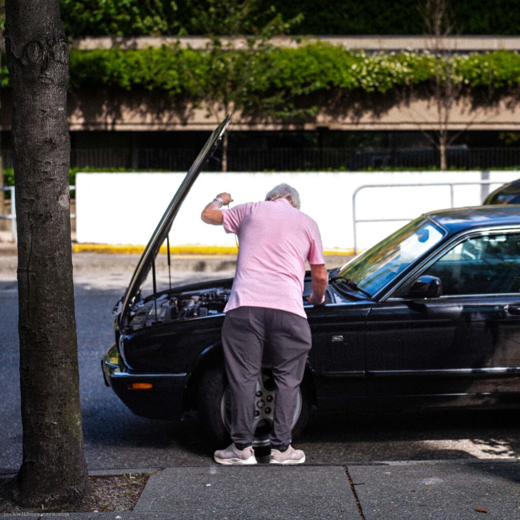 Man in pink shirt standing beside black car at curb, lowering or closing the open hood. Parking meter and trees visible.