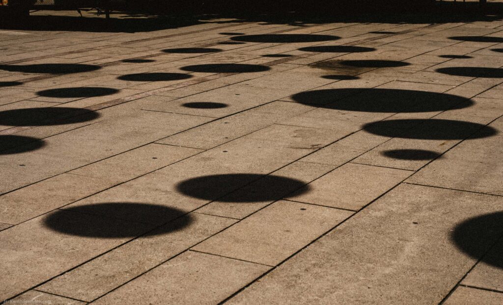 Multiple circular shadows on light stone plaza surface, slightly wider framing. Feet of a person visible at upper left edge.