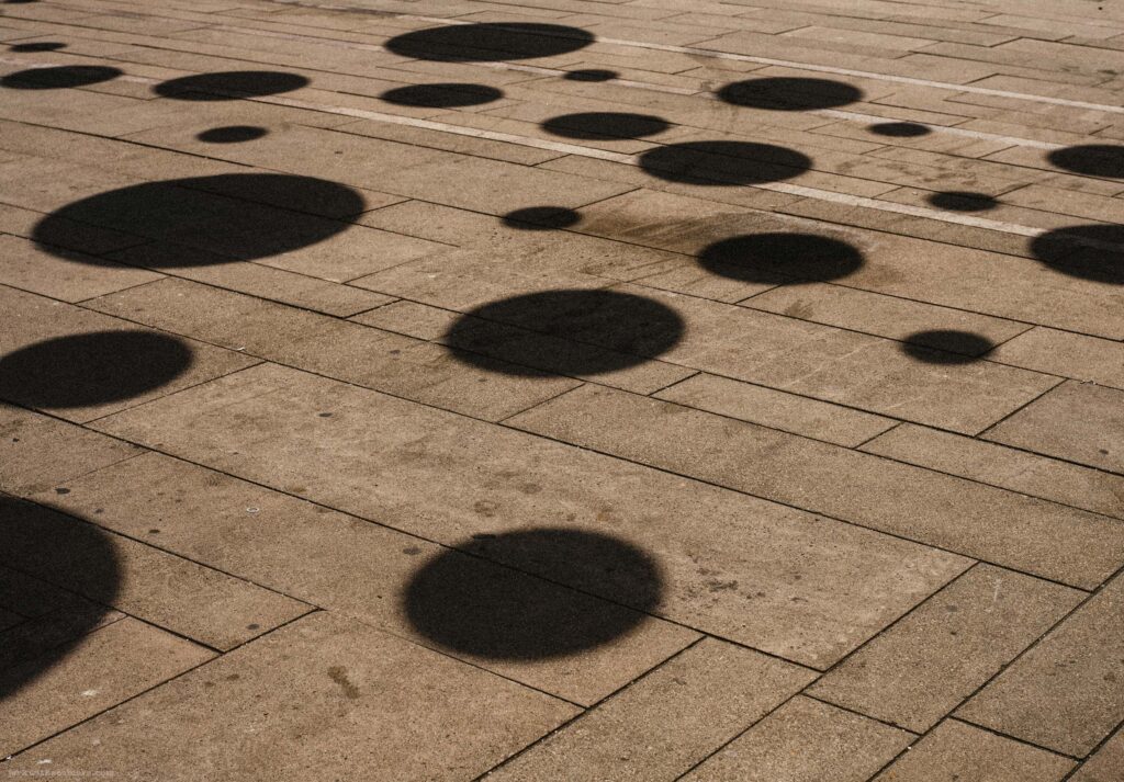 Widest view of circular shadows on light stone plaza paving. More of the surrounding surface visible. Small red object at centre.