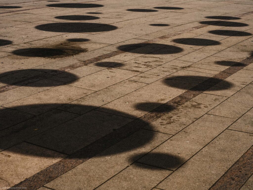 Circular shadows on stone plaza paving with feet and lower legs of a person visible at the upper left. Larger circular shapes dominate the frame.
