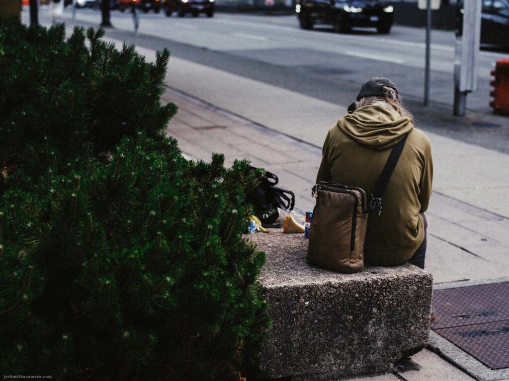 wearing olive jacket beside a conifer with a street and cars visible. Rotated 90 degrees.