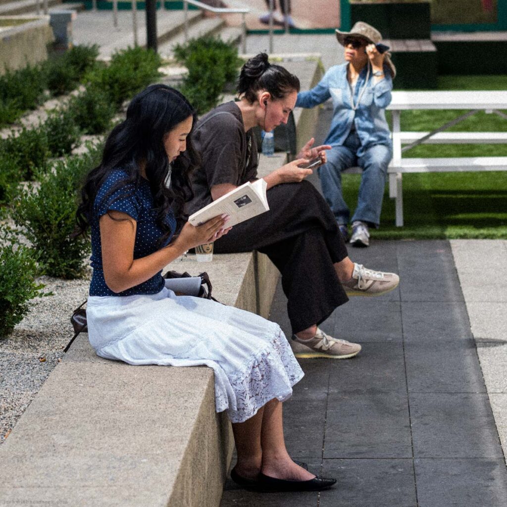 Three people sitting on a low stone ledge outside Simons. Two look at phones. Simons advertising banners and white sculptural sphere visible.