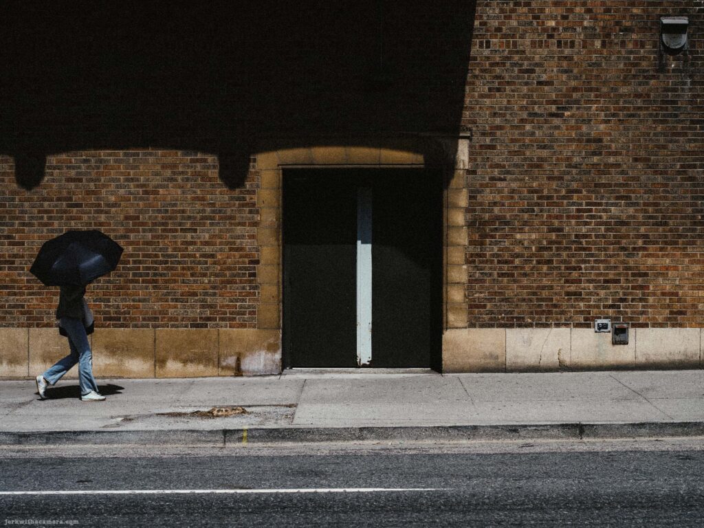 Person with large black umbrella walking past red brick building double door. No parking sign post visible at left.
